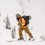 man tourist in bright yellow suit with skis and trekking poles walks up the hill against the backdrop of snow-covered evergreen trees