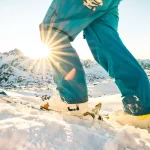 Legs of professional skier at sunset on relax moment in french alps ski resort - Winter sport concept with adventure guy on mountain top ready to ride down - Side view point with azure vintage filter